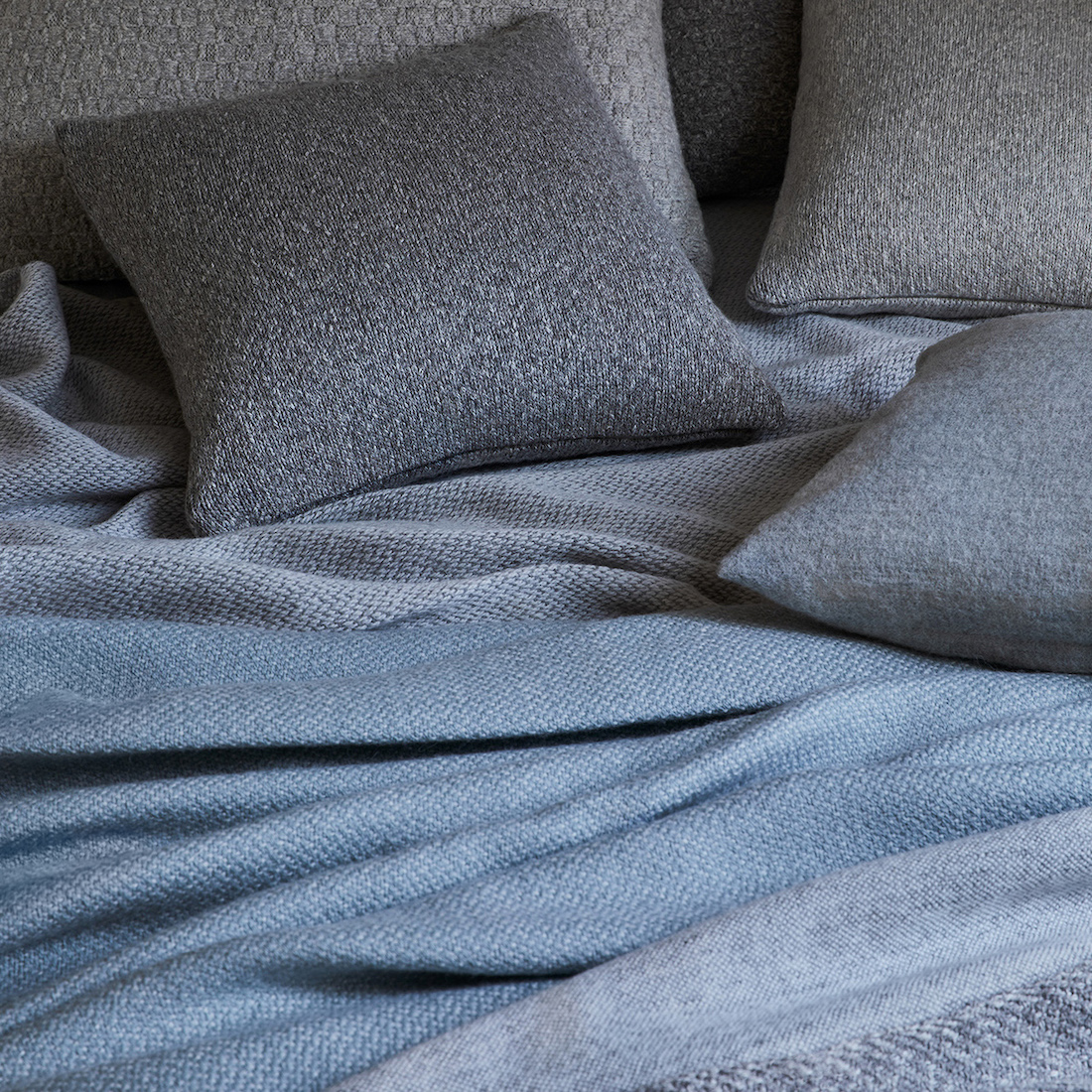 Pled dwustronny baby alpaca z kaszmirem i wełną merynosa w kolorze Ocean i Grey Mist, pled gładki Grey Mist, poduszki dekoracyjne baby alpaca i bawełna organiczna w kolorze Natural Silver i Natural Grey i poduszka 100% baby alpaca Classic Silver. 